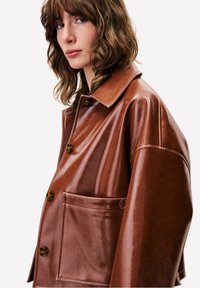 Vrouw met golvend bruin haar, die een glanzende bruine leren jas met grote knopen en een zichtbare voorkantzak draagt, lichtjes naar links gekeerd.