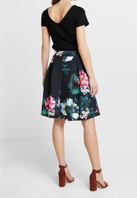 Černá květinová sukně s barevnými růžovými a zelenými vzory, spárovaná s přiléhavým černým svetrem s jemným žebrováním. Model nosí hnědé sandály na blokovém podpatku.