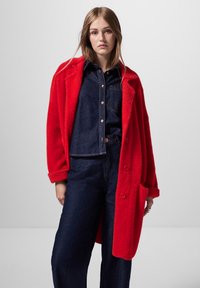 Röd plyschrock med oversized passform och uppvikt ärmar, prydd med stora knappar och två framfickor, bärs över en mörk denimtopp och byxor.