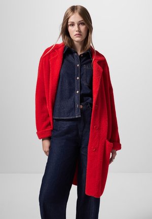 Rode pluche jas met een oversized pasvorm en opgerolde mouwen, met grote knopen en twee voorkruiden, gedragen over een donkerdenim top en broek.