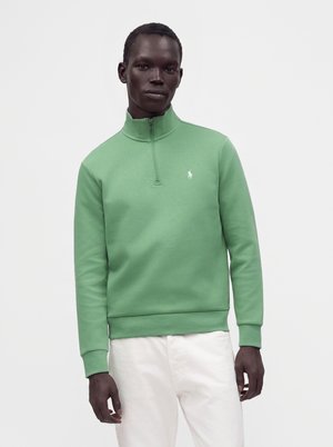 Mand iført en lysegrøn pullover med kvart lynlås og en lille hvid logo på brystet og hvide bukser, stående mod en ensfarvet baggrund.