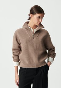Pull-over en tricot côtelé beige avec un col zippé mi-hauteur, présentant un design en V et une coupe décontractée, superposé sur une chemise blanche à col.