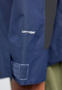 Blå vandtæt jakkeærme med "DRYVENT" logo i hvid broderi, med en struktureret velcro-rem til justeringer.