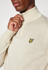 Cream-colored, half-zip sweater with a textured knit. Features a yellow embroidered logo on the chest and fitted ribbed cuffs.