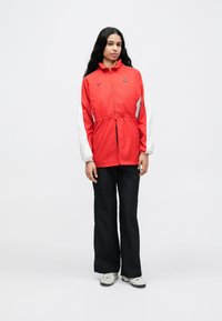 Jachetă ușoară roșie cu mâneci albe, prevăzută cu fermoar, talie elastică și logo Nike. Asortată cu pantaloni negri și adidași gri.