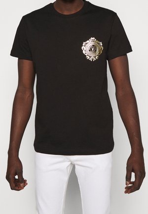 Man draagt een zwart T-shirt met korte mouwen met een goudkleurig, sierlijk cirkelvormig embleem op de linkerborst en witte broek, tegen een effen achtergrond.