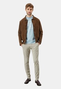 Giacca in suede marrone sopra una felpa grafica azzurro chiaro, abbinata a pantaloni beige chiaro e mocassini scuri, in piedi con le mani nelle tasche.