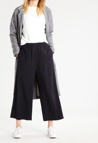 Grå rutig lång kappa över en vit topp, kombinerad med svarta vida byxor och vita sneakers. Slen tyg, lös passform och elastisk midja.