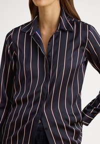 Sieviete tumsā zilā pogām aizdares kreklā ar vertikālām baltām un sarkanām svītrām, valkājot tumšas džinsas, stāv ar roku uz gurna.