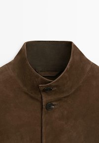 Braune Wildlederjacke mit hochstehendem Kragen und zwei schwarzen Knöpfen, die im Halsbereich geschlossen sind.