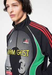 Ung person som har på seg en svart zip-up-jakke med røde striper, grønne paneler og "TEAM GEIST" i gul på forsiden sammen med Adidas-logo.