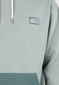 Haut à capuche vert clair avec un ourlet vert foncé. Présente un petit logo Oxbow brodé sur la poitrine et des cordons de serrage avec des embouts en métal.
