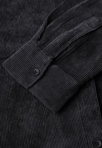 Black corduroy shirt detail focuses on textured fabric, with prominent vertical ribbing, a buttoned cuff, and smooth button accents.