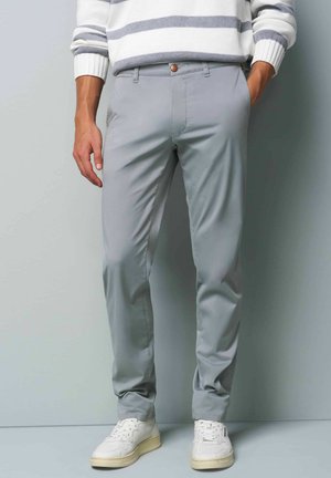 Helle graue Chinos mit glatter Textur, slim fit und einem einzelnen Knopf vorne. Kombiniert mit weißen Sneakern, stehend vor einem neutralen Hintergrund.