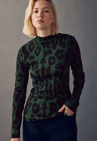 Hosszú ujjú, sötétzöld garbó fekete leopárdmintával, bordázott textúrával, karcsúsított szabással és klasszikus nyakkivágással, sötét farmerrel párosítva.