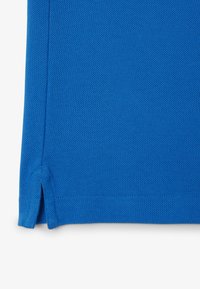 Prenda de color azul brillante con un tejido texturizado, que presenta aberturas laterales en el dobladillo y costuras reforzadas. Sin patrones visibles ni detalles de hardware.