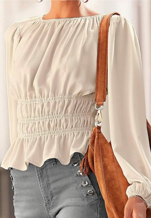 Mujer vistiendo una blusa de crema con fruncidos, pantalones grises de cintura alta con botones, y llevando un bolso de hombro de ante marrón con detalle de borlas.