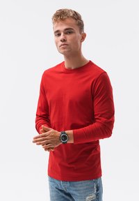 Langärmliges rotes T-Shirt aus einer Baumwollmischung, mit Rundhalsausschnitt und glatter Textur, kombiniert mit distressed blauen Jeans und einer schwarzen Uhr.