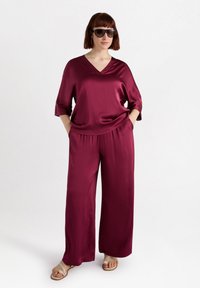 Burgundy satin topp med V-hals och vida ärmar, parat med matchande vida byxor; modellen bär solglasögon och sandaler.