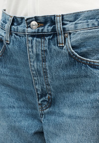 Κοντινό πλάνο στη μέση μπλε τζιν παντελονιού με μεταλλικό κουμπί, θηλιές για ζώνη και ορατή ραφή στην μπροστινή τσέπη.