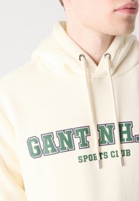 Krémová mikina s kapucňou a predným logom "GANT NH. SPORTS CLUB" v tmavozelenej a purpurovej farbe, so striebornými šnúrkami na sťahovanie.
