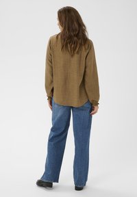 Braunes, strukturiertes Langarmhemd mit gerafften Manschetten, kombiniert mit lockeren blauen Jeans und schwarzen Schuhen, von hinten vor hellem Hintergrund gesehen.