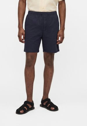 Mężczyzna noszący granatowe szorty z troczkiem, czarne sandały na paski oraz jasnobeżową koszulę z krótkim rękawem na białym tle.