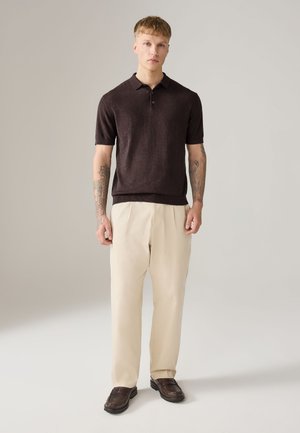 Fiatal férfi tetoválásokkal, sötétbarna rövid ujjú galléros pólót, krémszínű redőzött nadrágot és sötétbarna mokaszint visel, világos háttér előtt áll.