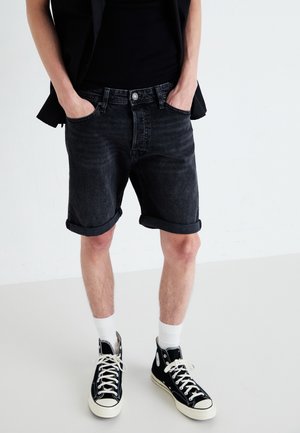 Person trägt hochgekrempelte schwarze Denimshorts, schwarze High-Top-Sneaker mit weißen Schnürsenkeln, weiße Socken und ein schwarzes ärmelloses Oberteil.