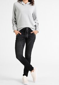 Sudadera gris claro con cordón, bordado floral en las mangas; combinada con jeans negros ajustados y zapatillas beige. Textura suave, diseño casual.