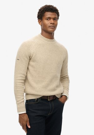 Suéter de punto color beige claro con cuello y puños acanalados, que presenta un patrón texturizado. Combinado con jeans de mezclilla oscura y un cinturón marrón.