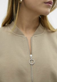 Veste beige zippée avec un col côtelé, dotée d'une fermeture éclair argentée et d'une languette de tirage circulaire. Le tissu semble doux avec une texture lisse.