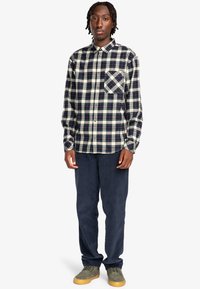 Camisa de franela a cuadros en navy, crema y burdeos, con cuello abotonado y un bolsillo en el pecho. Combinada con pantalones navy y zapatos de ante.
