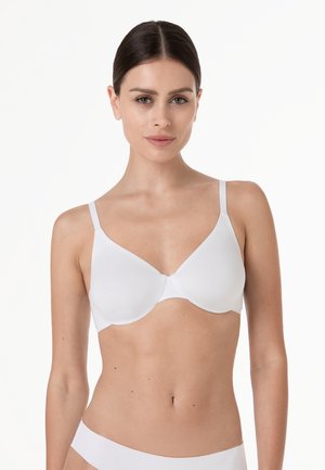 Donna con capelli scuri che indossa un reggiseno bianco con ferretto liscio e biancheria intima bianca abbinata su uno sfondo bianco semplice.
