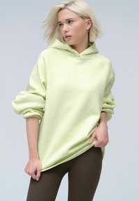 Haut à capuche oversize vert clair avec cordon de serrage, poignets côtelés et texture douce, associé à des leggings côtelés marron foncé.