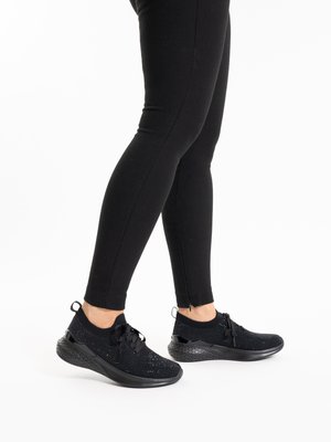 Personne portant un pantalon noir ajusté et des baskets de sport en maille noire avec lacets, debout sur un fond blanc.