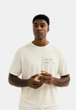 Ung mand i hvid t-shirt, der holder en gul padelbold og kigger let til siden mod en ensfarvet hvid baggrund.