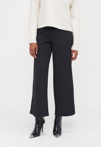 Schwarz-weiße, weit geschnittene Hose mit vertikalen Streifen, aus einem glatten Stoff gefertigt. Kombiniert mit spitzen, schwarzen Stiefeletten für einen Kontrast.