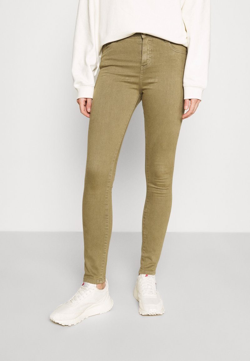 Pantalon slim vert olive porté par une personne debout, associé à des baskets blanches épaisses et un top blanc à manches longues.