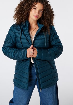 Femme aux cheveux bouclés portant une veste matelassée bleu sarcelle foncé sur un haut blanc et un jean bleu, tenant la fermeture éclair de la veste au niveau de la poitrine.