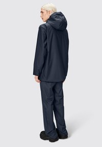 Giacca impermeabile da marina con cappuccio, abbinata a pantaloni coordinati. Tessuto liscio, vestibilità comoda e design minimalista. Calzature nere texturizzate.