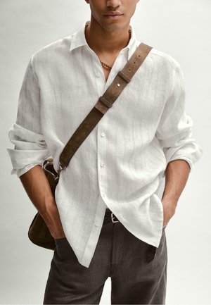 Mężczyzna ubrany w białą lnianą koszulę z podwiniętymi rękawami, ciemne spodnie, brązową skórzaną torbę na ramię i widoczny naszyjnik z koralików.