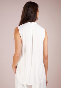 Top blanco sin mangas con cuello alto, que presenta un pliegue en la parte trasera y un pequeño detalle de logotipo cerca del cuello. Textura de tela suave y lisa.