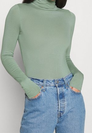 Vrouw die een nauwsluitende, saliegroene coltrui met lange mouwen draagt, ingestopt in een lichtblauwe, hooggelede spijkerbroek, met haar handen in de zakken.