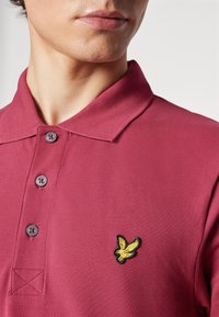 Burgundská polo košeľa s textúrovanou látkou, s tromi sivými gombíkmi a čierne orámovaným žltým výšivkou vtáka na ľavej hrudi.
