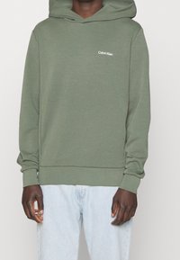 Zelená mikina s kapucí vyrobená z měkké látky. Obsahuje malé bílé logo Calvin Klein na hrudi a vroubkované manžety a spodní lem.