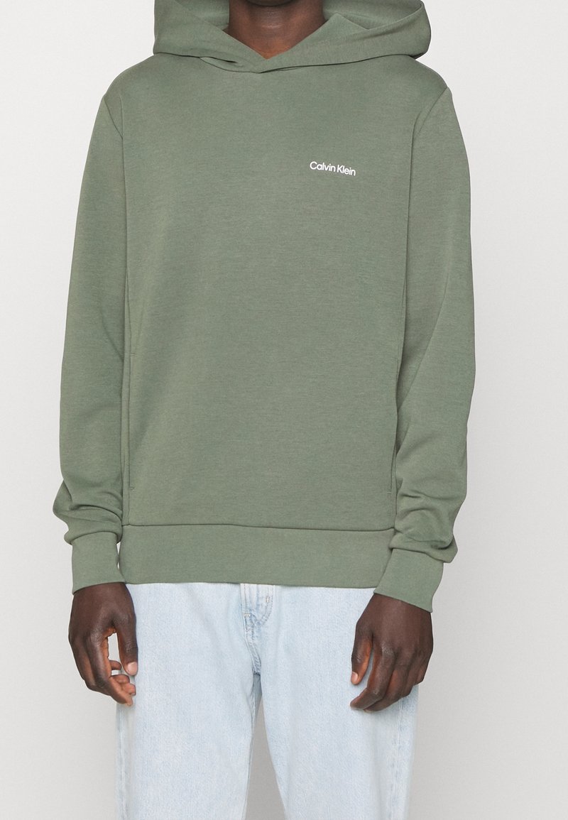 Zelená mikina s kapucí vyrobená z měkké látky. Obsahuje malé bílé logo Calvin Klein na hrudi a vroubkované manžety a spodní lem.