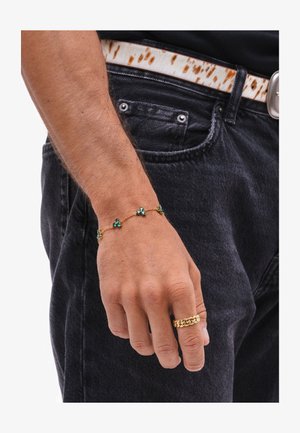 Mână masculină purtând o brățară din lanț de aur cu pietre verzi și un inel masiv din lanț de aur, odihnindu-se lângă blugi negri din denim cu o curea albă și maro.