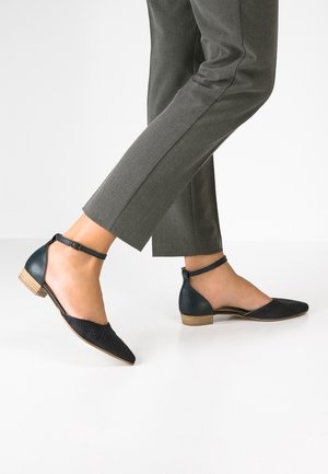 Zapatos negros de tacón bajo con tiras en el tobillo y punta afilada, combinados con pantalones grises recortados sobre un fondo claro.