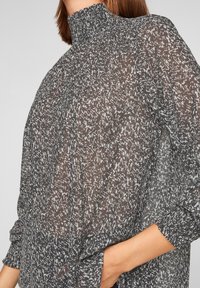Kvinna klädd i en höghalsskjorta med lång ärm, genomskinlig mönstrad blus med handen instucken i byxfickan framför en neutral bakgrund.